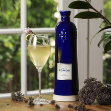 Load image into Gallery viewer, Komos Anejo Cristalino Tequila in its deep blue artisan ceramic bottle next to a pale green cocktail on a wooden surface with greenery in the background