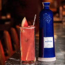 Load image into Gallery viewer, Komos Anejo Cristalino Tequila in its deep blue artisan ceramic bottle next to a pink cocktail with watermelon garnish