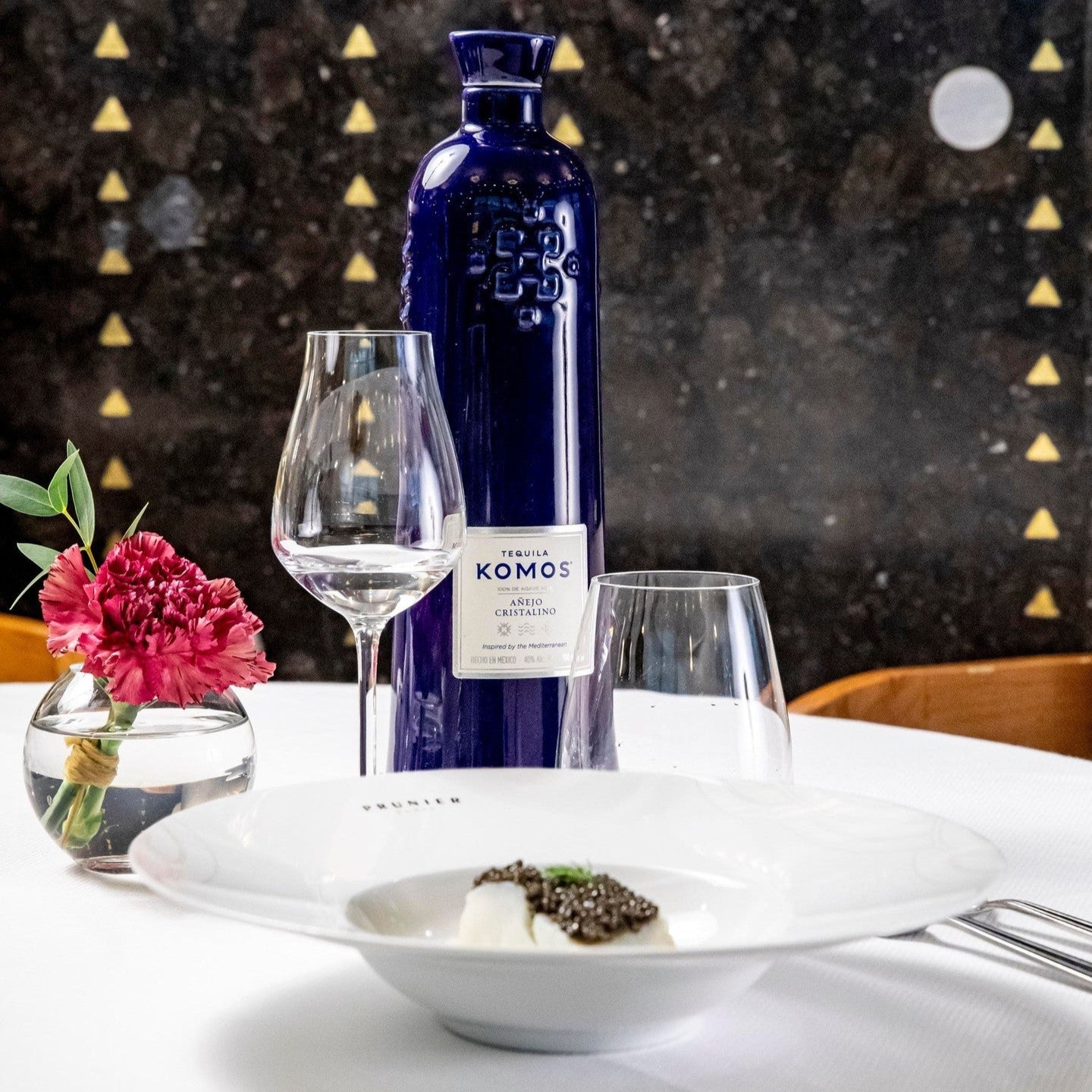 Load image into Gallery viewer, Komos Anejo Cristalino Tequila in its deep blue artisan ceramic bottle set on a dinner table with glasses and flower decoration