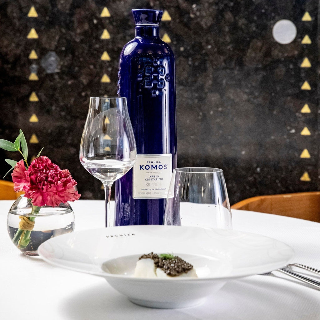 Komos Anejo Cristalino Tequila in its deep blue artisan ceramic bottle set on a dinner table with glasses and flower decoration