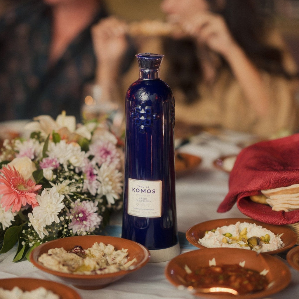 Komos Anejo Cristalino Tequila in its deep blue artisan ceramic bottle set on a table with food and flowers