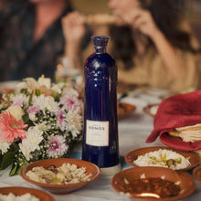 Load image into Gallery viewer, Komos Anejo Cristalino Tequila in its deep blue artisan ceramic bottle set on a table with food and flowers