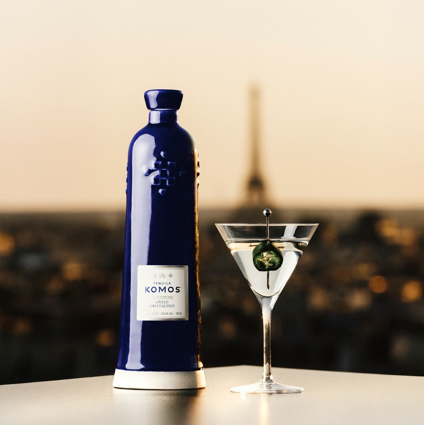 Load image into Gallery viewer, Komos Añejo Cristalino Tequila with a martini glass containing a olive on a blurred Paris city background