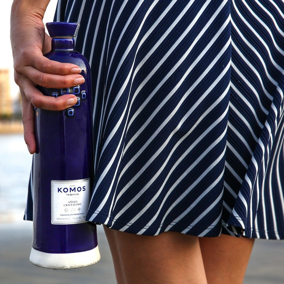 Load image into Gallery viewer, Komos Anejo Cristalino Tequila in its deep blue artisan ceramic bottle being held in a womans hand with harbour background