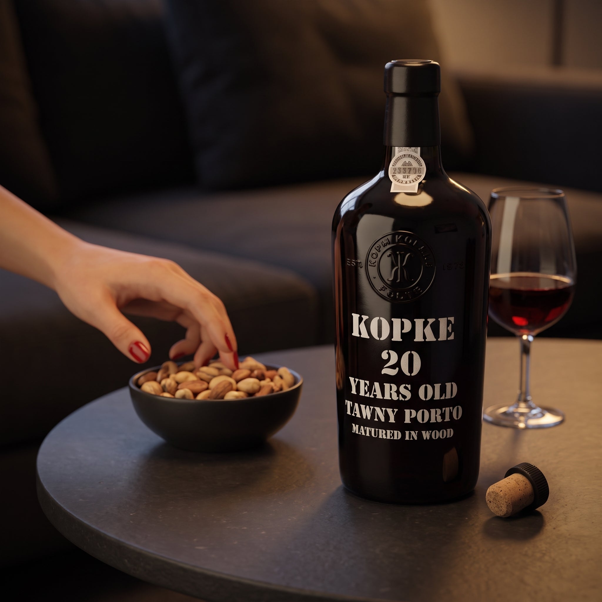 Load image into Gallery viewer, A dark bottle of Kopke 20 Year Old Tawny Port wine stands on a round table beside a glass of red wine, a small bowl of nuts, and a cork, with a person's hand reaching toward the nuts in a dimly lit living room setting.