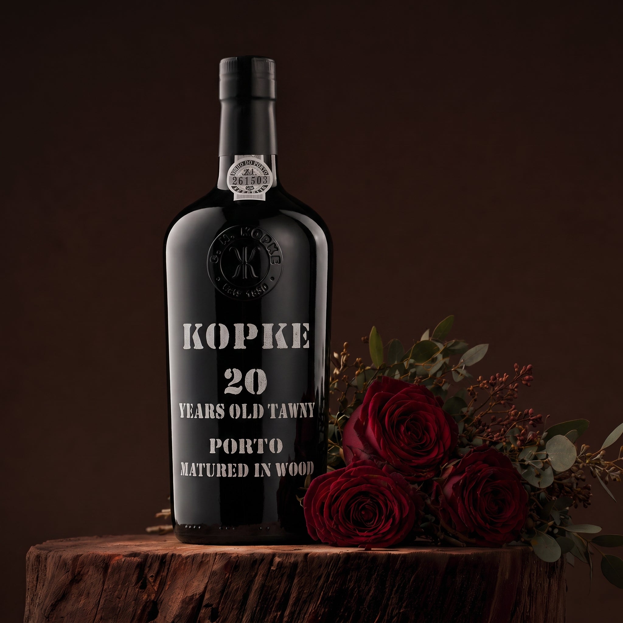 Load image into Gallery viewer, A Kopke 20 Year Old Tawny Port wine bottle with white lettering, styled on a rustic wooden stump next to three red roses against a dark brown background.