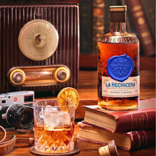 Load image into Gallery viewer, Bottle of La Hechicera fine aged rum with a glass of rum and ice, vintage radio, and books on a wooden table.
