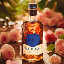Load image into Gallery viewer, Bottle of La Hechicera fine aged rum with a blue seal on a wooden surface surrounded by pink roses.
