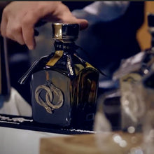 Load image into Gallery viewer, Hand holding a bottle of La-Hora-Azul-Blue-Hour-Reposado-Tequila with intricate emblem design, on a blurred background.