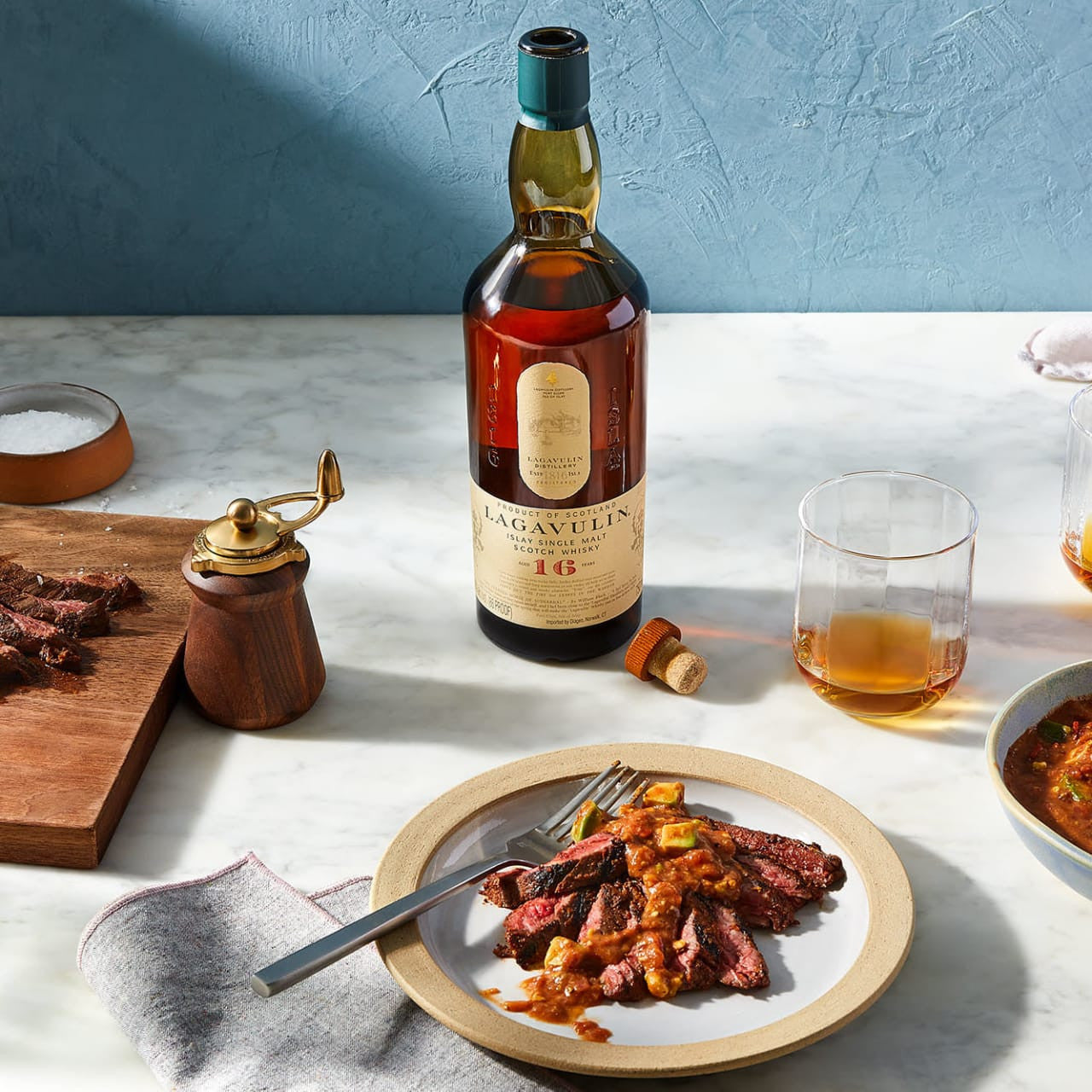 Load image into Gallery viewer, A bottle of Lagavulin 16YO Single Malt Whisky standing on a marble table with a plate of beautifully cut meat