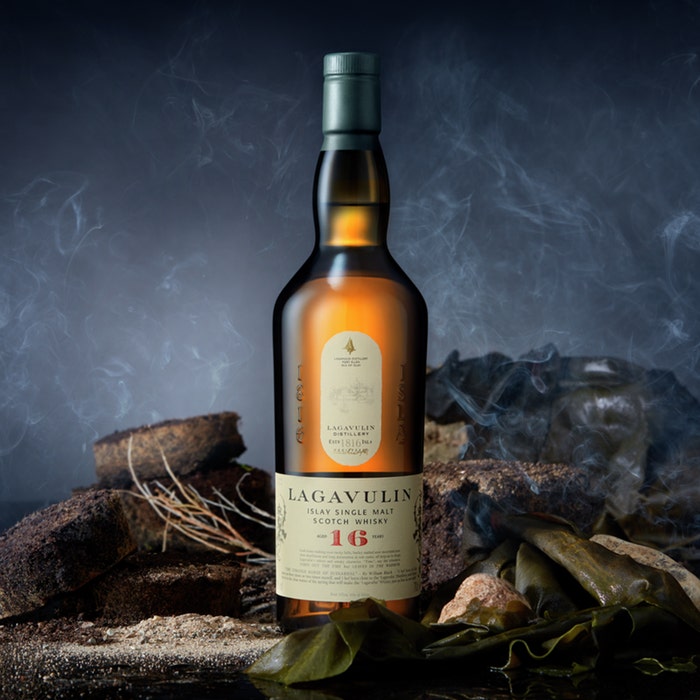 Load image into Gallery viewer, A bottle of Lagavulin 16 year old Single Malt Whisky standing on some rocks with a smoky background