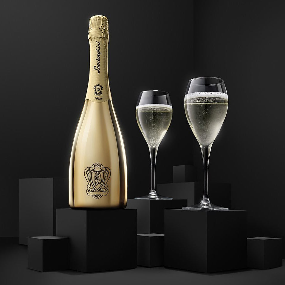 Load image into Gallery viewer, A gold-toned Lamborghini Brut Vino Spumante Gold bottle stands on a black pedestal beside two filled champagne flutes against a dark studio background.