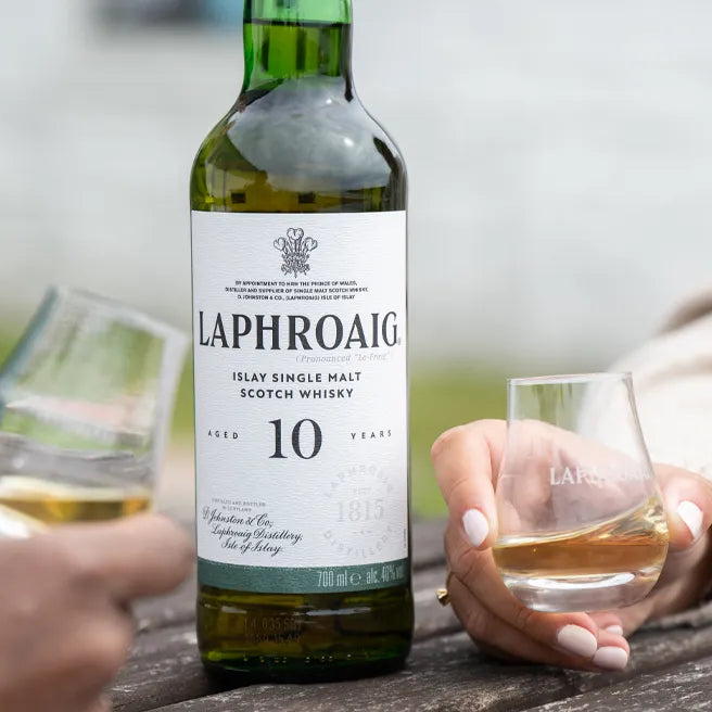 Bottle of Laphroaig 10 year old single malt whisky with two glasses being held by people outdoors.