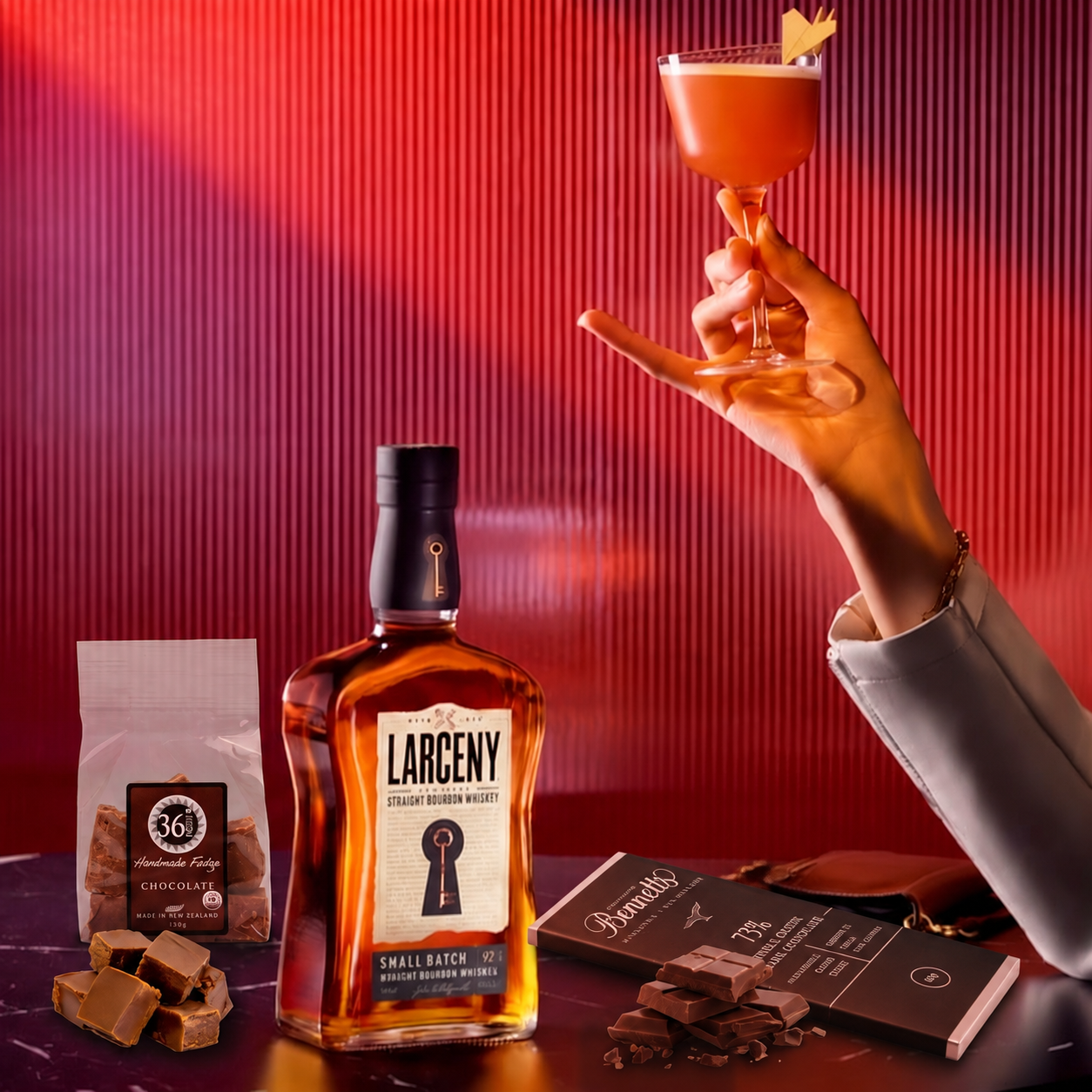 Load image into Gallery viewer, A bottle of Larceny Kentucky Straight Bourbon Whiskey is displayed on a dark table alongside a bag of 36 Degrees chocolate fudge, a bar of Bennett's 73% dark chocolate, chocolates fudge pieces and chocolate pieces, while a woman's hand holds a pink cocktail in a coupe glass against a red ribbed background.