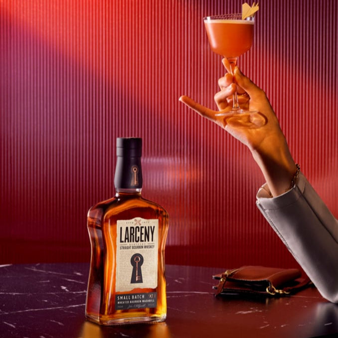 Load image into Gallery viewer, Larceny whiskey bottle with a hand holding a cocktail against a red background