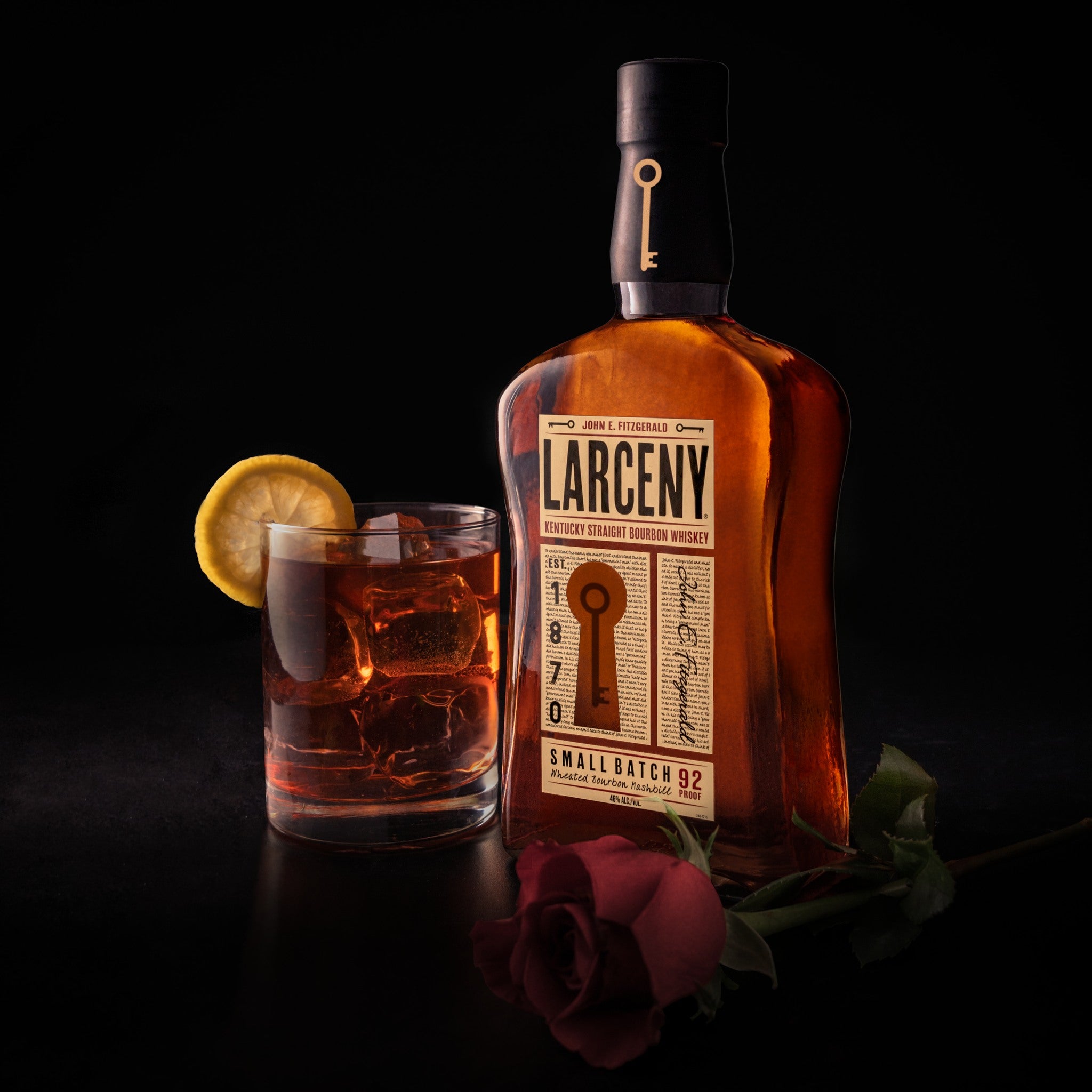 Load image into Gallery viewer, A bottle of Larceny Kentucky Straight Bourbon Whiskey and a tumble of whiskey with a lemon wedge standing on a black  table