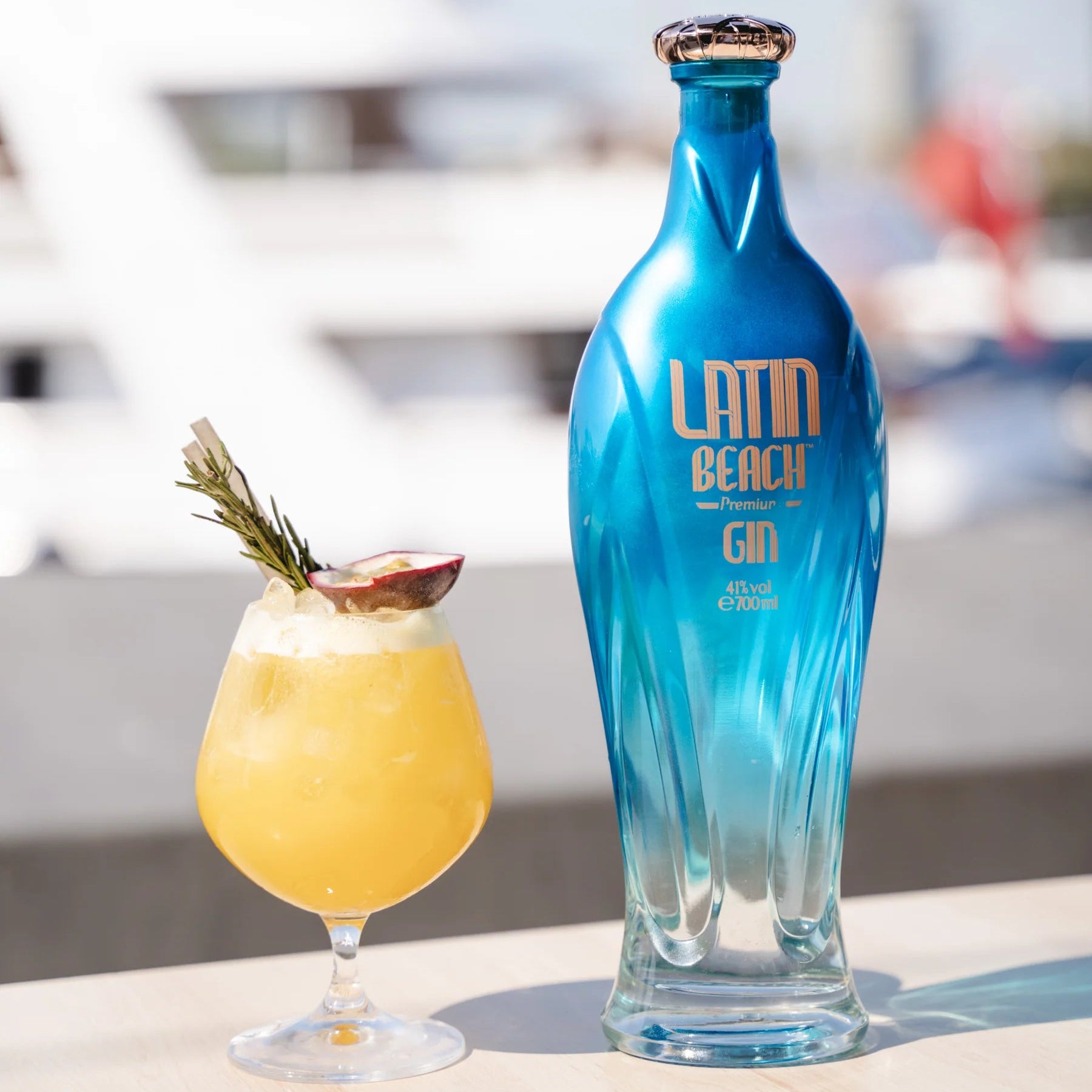 Load image into Gallery viewer, A blue bottle of Latin Beach Gin next to a cocktail with a blurred marina background