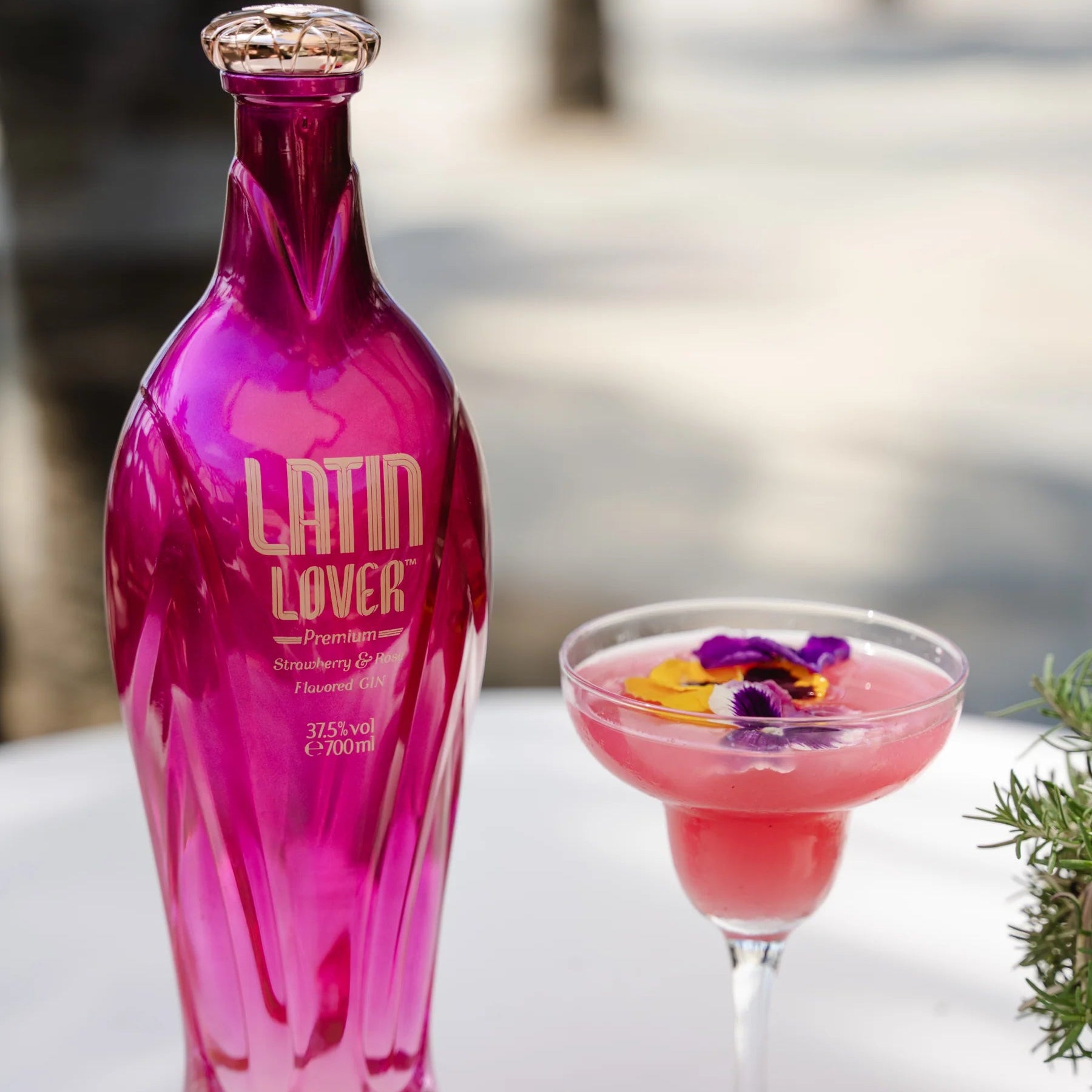 Load image into Gallery viewer, Pink Latin Lover Gin bottle next to a pink cocktail with flowers on a blurred outdoor background