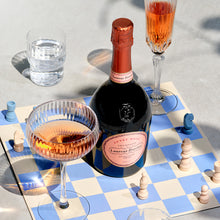 Load image into Gallery viewer, A bottle of pink champagne on a blu and white chessboard with some glasses 