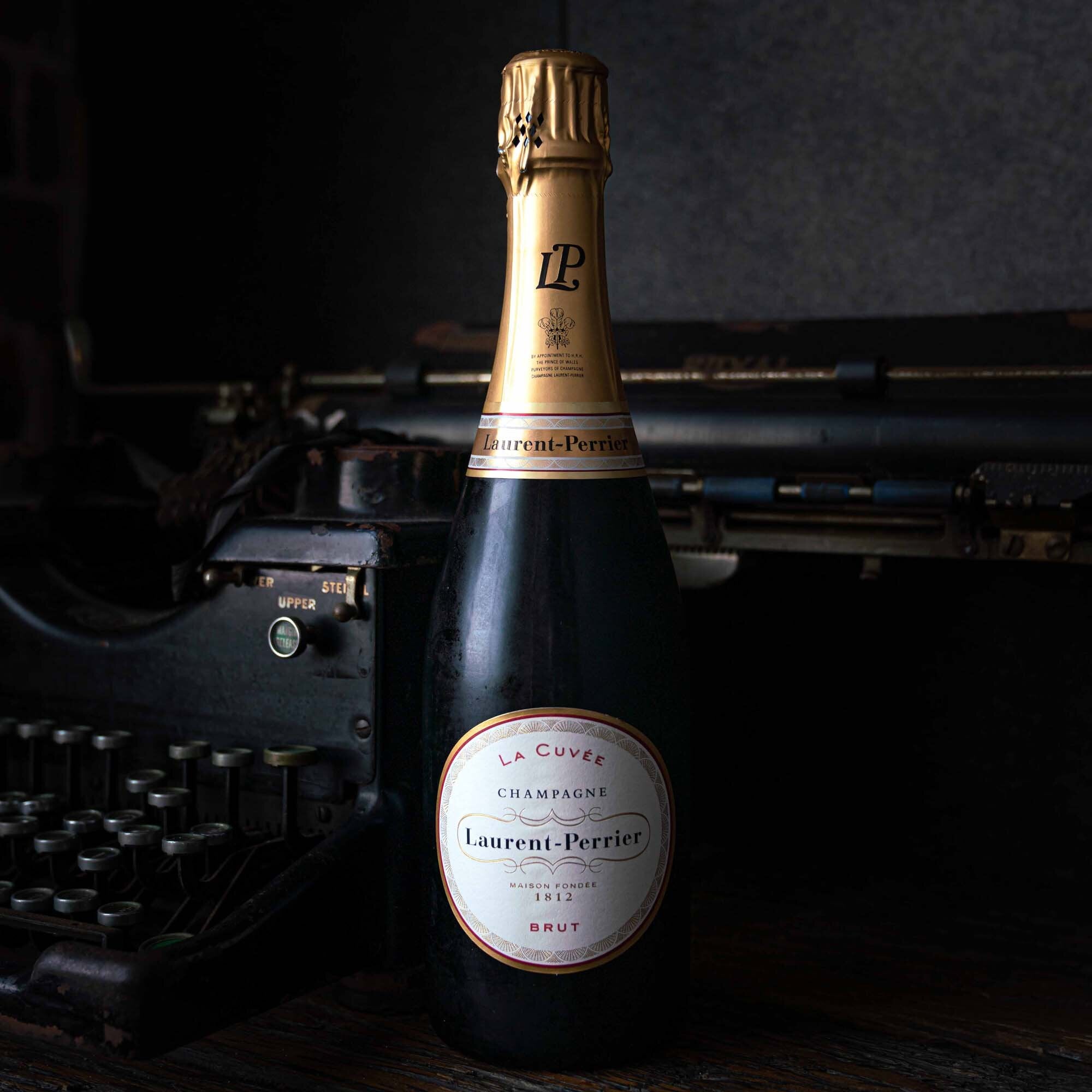 Load image into Gallery viewer, A bottle of Laurent-Perrier La Cuvée Brut Champagne and an old typewriter on a dark background