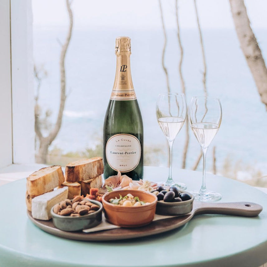 Bottle of Laurent-Perrier champagne with a charcuterie board on a table by a window.