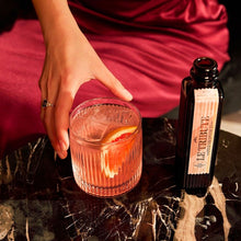 Load image into Gallery viewer, A woman in a red dress holding a cocktail glass with a bottle Le Tribute Pink Grapefruit Tonic Water on a dark surface.