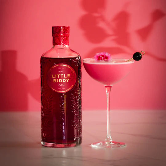 Bottle of Pink Little Biddy Gin next to a pink cocktail with a cherry on a pink background