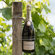 Load image into Gallery viewer, Bottle of Louis Roederer champagne tied to a wooden post with green foliage in the background