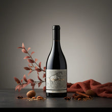 Load image into Gallery viewer, A bottle of red wine and nuts and flowers on a table.