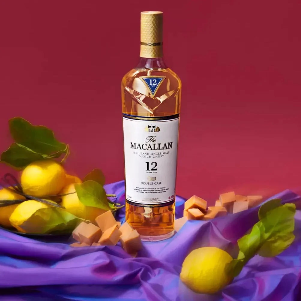 Load image into Gallery viewer, A bottle of whisky and lemons and fudges on a silky, purple tablecloth with a pink background.