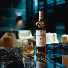 Load image into Gallery viewer, Macallan 15 year old whiskey bottle with a glass on a dark marble surface in a dimly lit room.