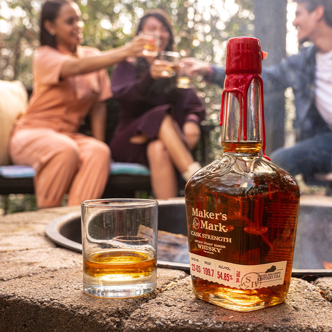 Load image into Gallery viewer, Maker's Mark whiskey bottle and glass on a stone surface with people in the background