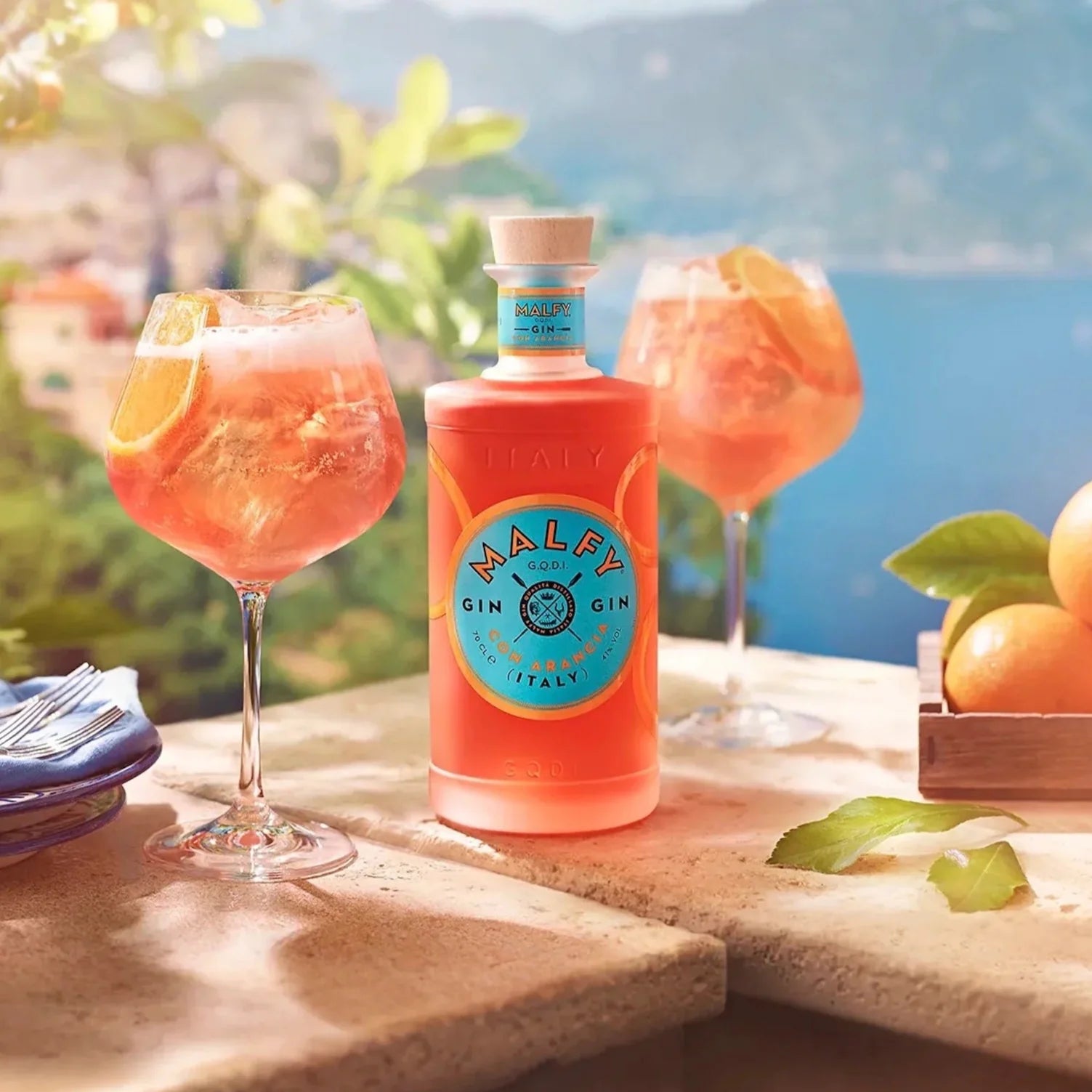 Load image into Gallery viewer, A bottle of pink gin with a couple of gin glasses standing on a stone table with a beautiful background of the sea and mountains in Italy.