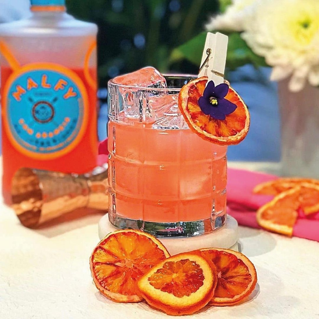 Load image into Gallery viewer, A bottle of Malfy Arancia Orange gin and a cocktail with orange slices and flower garnish on a table.