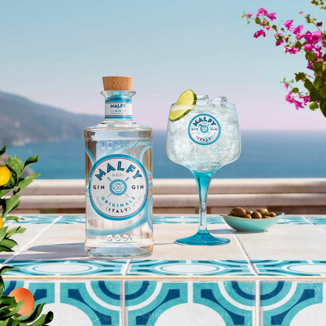 Load image into Gallery viewer, Bottle of Malfy Originale gin and a gin glass with a lime garnish on a tiled table by the sea.