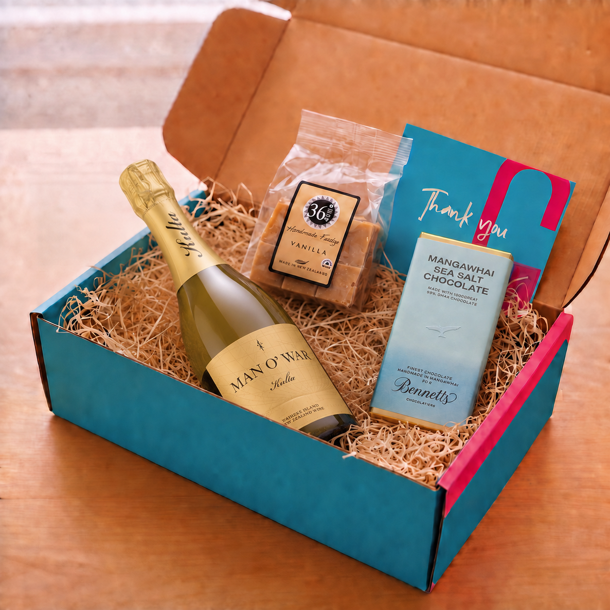 Load image into Gallery viewer, An open gift box filled with wood-wool packing material on a wooden surface, containing a bottle of Man O' War Kulta Tulia Blanc de Blac Sparkling, a bag of 36 Degrees Vanilla fudge, a blue and pink Bennett's Mangawhai Sea Salt Chocolate bar, and a blue thank-you card.