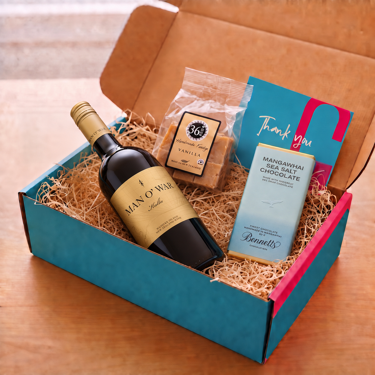 Load image into Gallery viewer, A curated gift box set on a wooden table, featuring a bottle of Man O' War Kulta 'Tytti' Bordeaux Blend red wine, a bag of 36 Degrees Vanilla vanilla fudge, a bar of Bennett's Mangawhai Sea Salt Chocolate, and a teal gift card, arranged in a COVE 27 teal and pink box with wood-wool.
