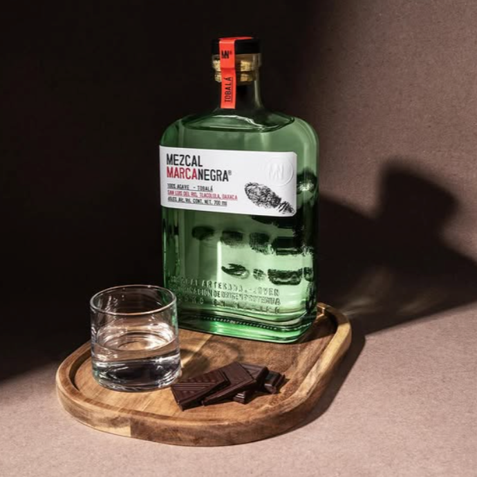 Load image into Gallery viewer, Green bottle of Mezcal Marca Negra with a glass and chocolate on a wooden tray against a dark background