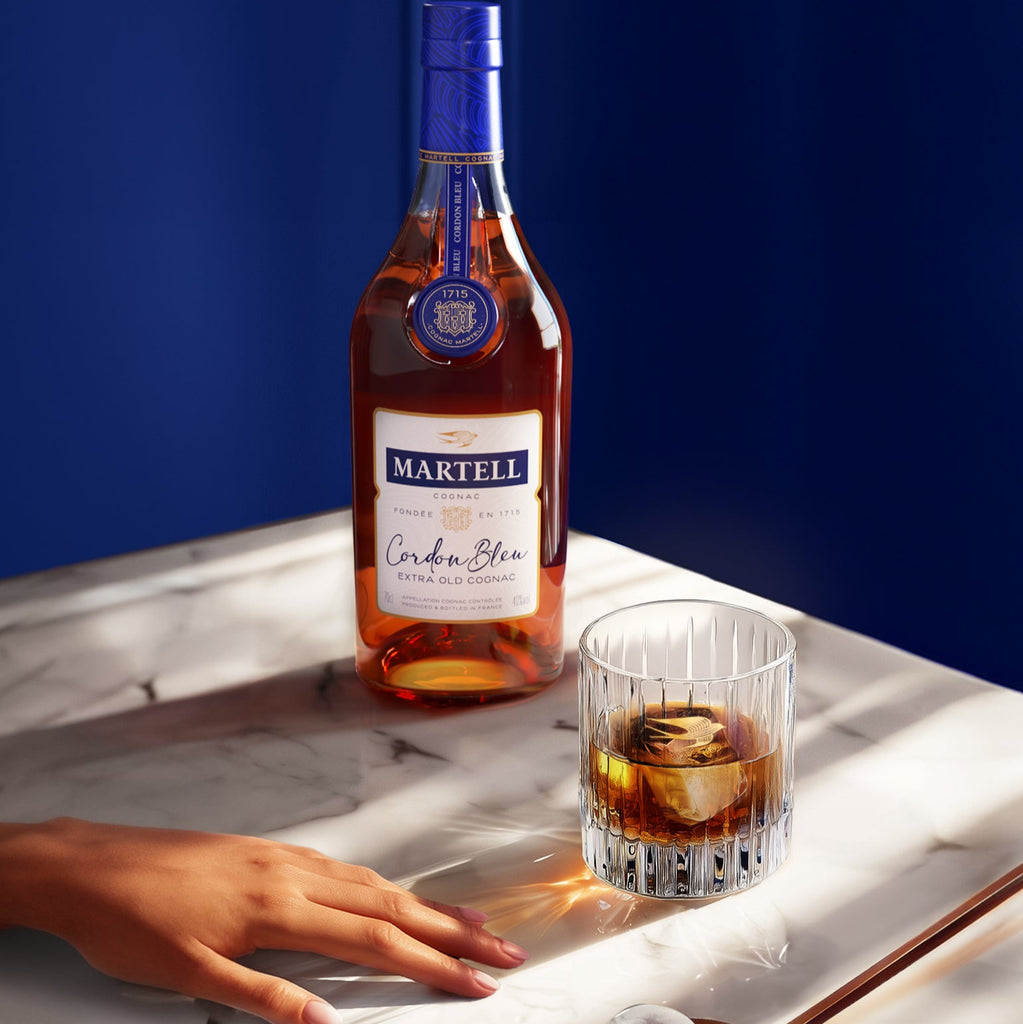 A bottle of Martell Cordon Bleu Cognac and a hand reaching for a glass of cognac with ice in the glass
