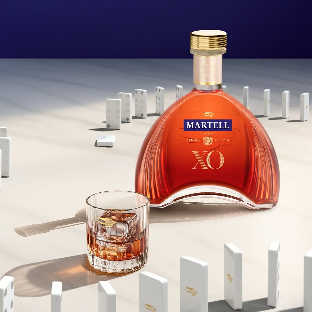 Load image into Gallery viewer, A bottle of Martell XO cognac with a gold cap stands on a white surface beside a clear rocks glass filled with an amber drink. White dominoes are arranged around the scene against a dark blue background.