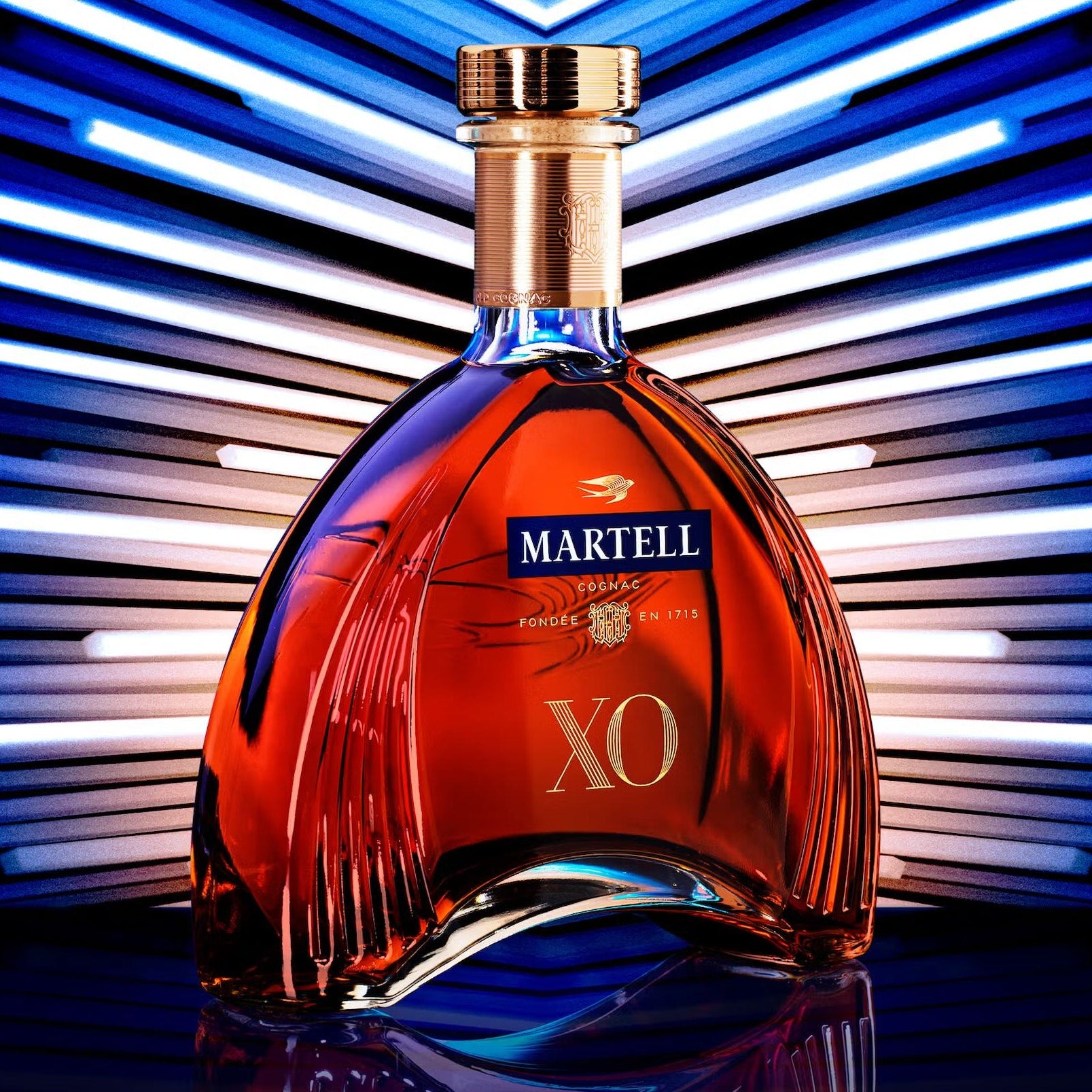 Load image into Gallery viewer, A bottle of Martell XO cognac centered against a stylised background of blue and white light streaks. The bottle is made of clear glass showing the amber-red liquid inside, with a gold cap and a blue and gold Martell XO label.