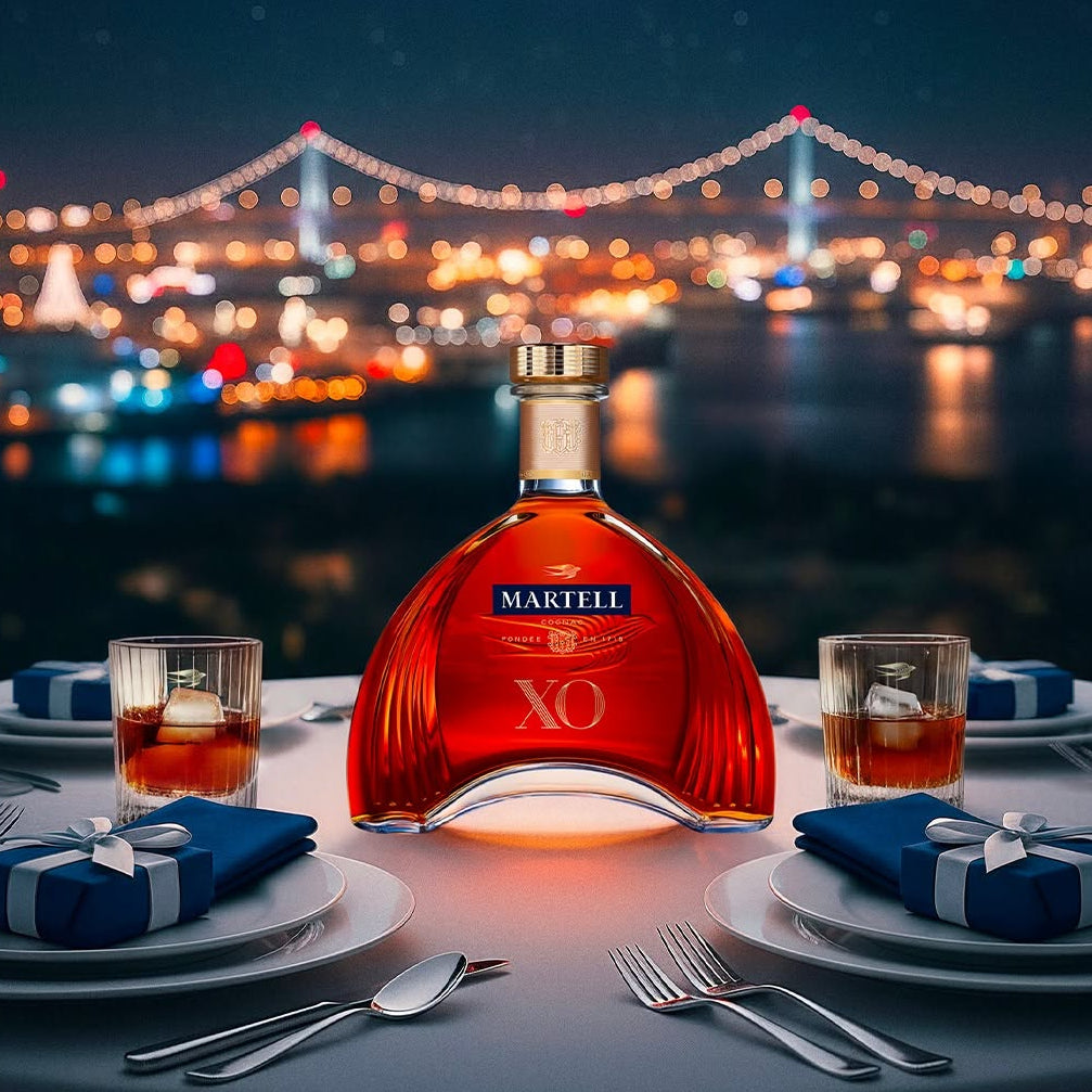 Load image into Gallery viewer, A bottle of Martell XO cognac is centered on a dining table at night, with two glasses, plates, folded blue napkins with white ribbons, and a blurred city bridge in the background.