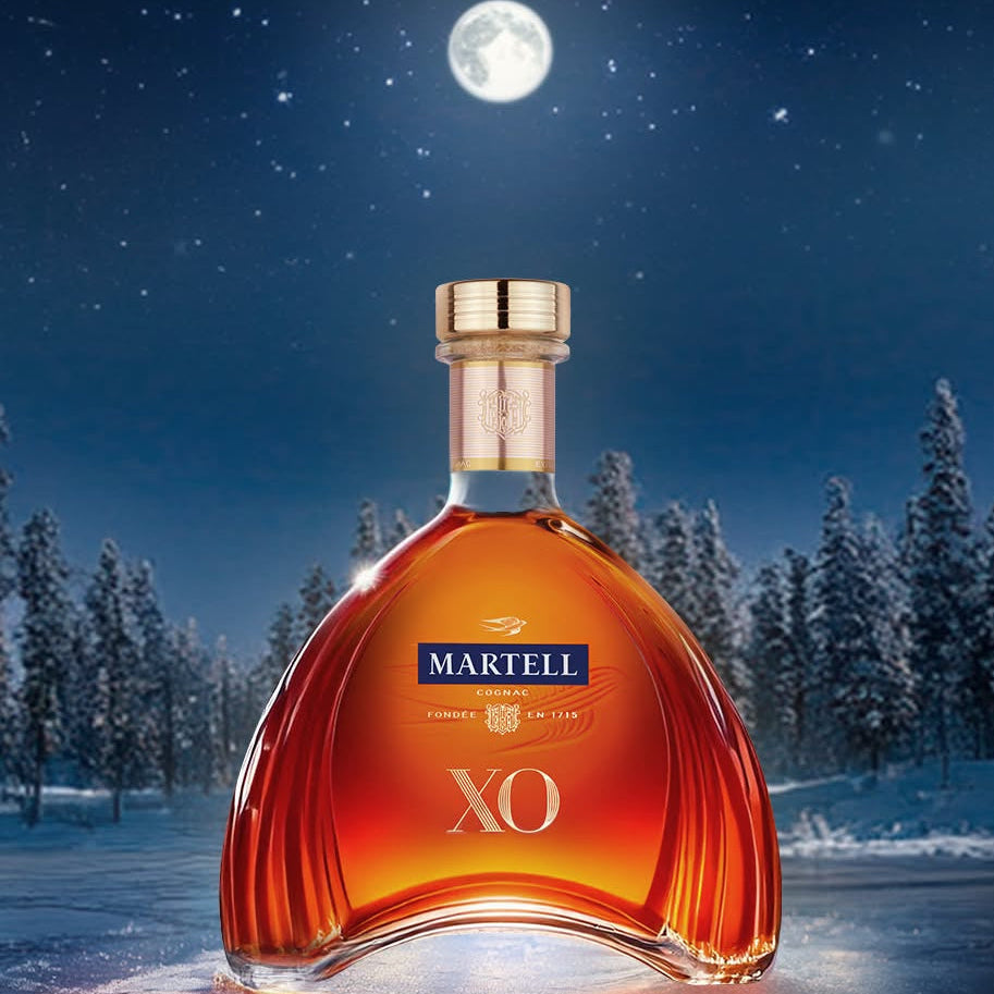 Load image into Gallery viewer, A Martell XO cognac bottle stands in a snowy night landscape with pine trees and a full moon in the background. The bottle is filled with amber liquor and has a gold cap, with the Martell logo and XO text visible on the front.