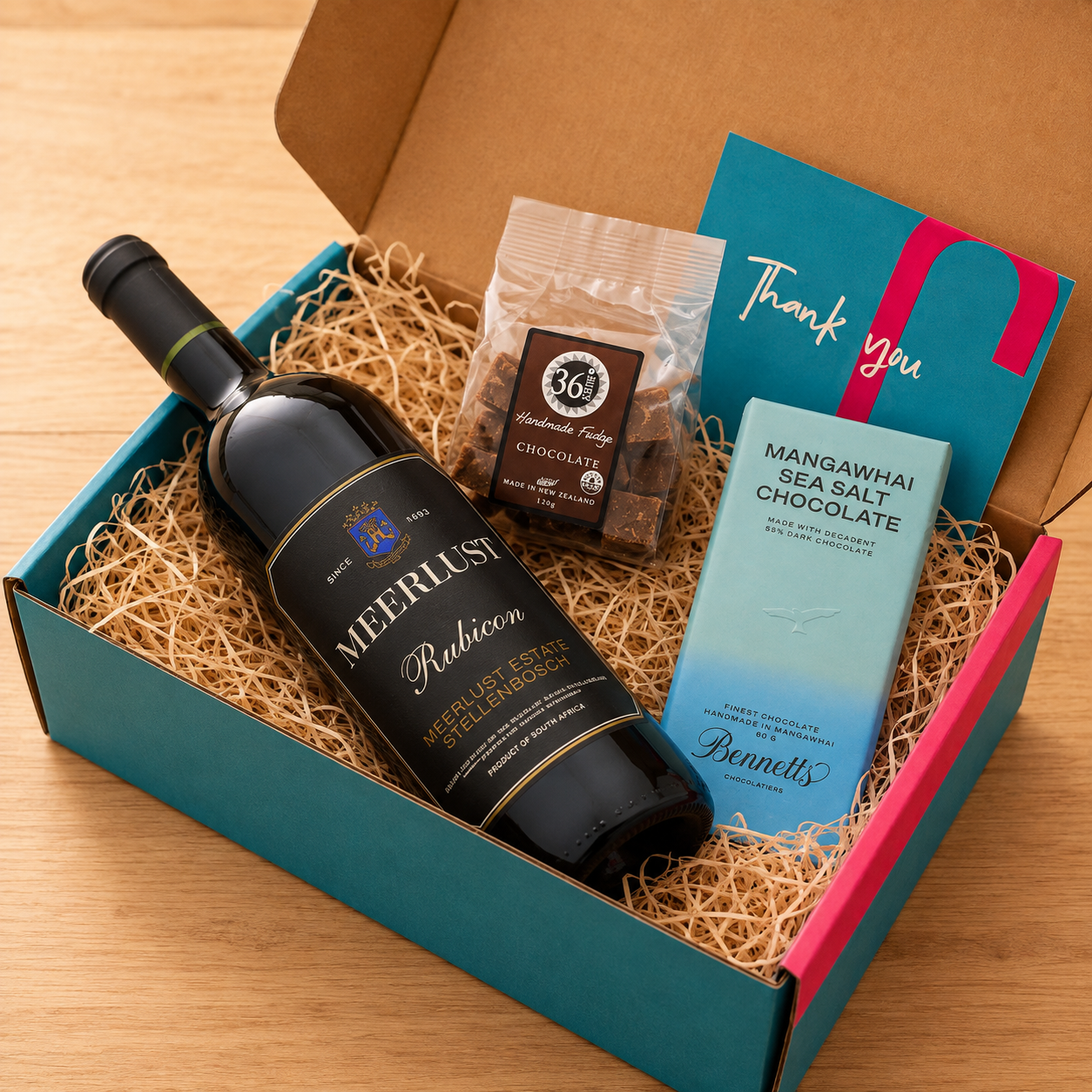 Load image into Gallery viewer, A curated gift box set on a wooden table, featuring a bottle of Meerlust Estate Rubicon red wine, a bag of 36 Degrees chocolate fudge, a blue box of Mangawhai Sea Salt Chocolate by Bennetts, and a teal thank you card inside a teal and pink gift box with shredded paper.