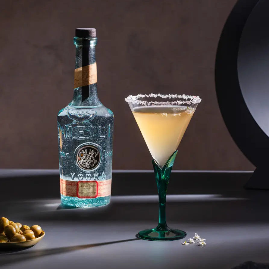 Load image into Gallery viewer, A clear glass Meili Vodka bottle with a black cap and gold label details stands beside a martini-style cocktail glass filled with pale yellow drink and a sugar-rimmed edge, set on a dark tabletop.