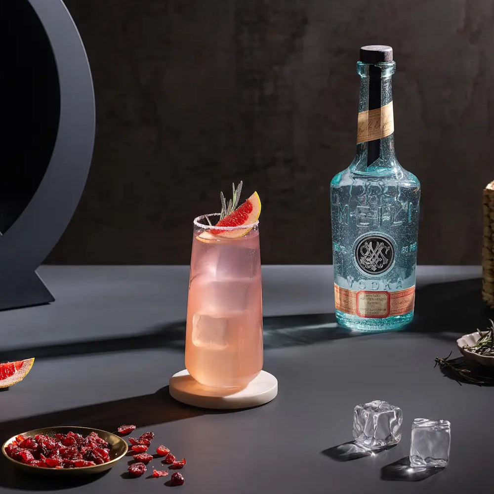 Load image into Gallery viewer, A styled cocktail scene on a dark tabletop with a pink drink in a tall glass, a Meili Vodka bottle, ice cubes, dried red berries, and grapefruit garnish against a dark studio background.