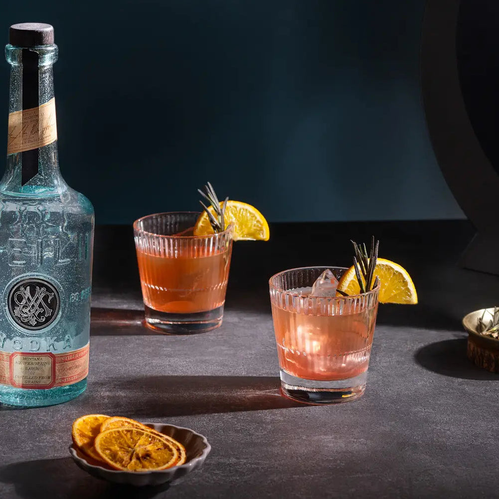 Load image into Gallery viewer, A bottle of Meili Vodka sits beside two rocks glasses filled with an orange-pink cocktail, garnished with orange slices and rosemary, on a dark tabletop with dried orange slices and a small dish of herbs.