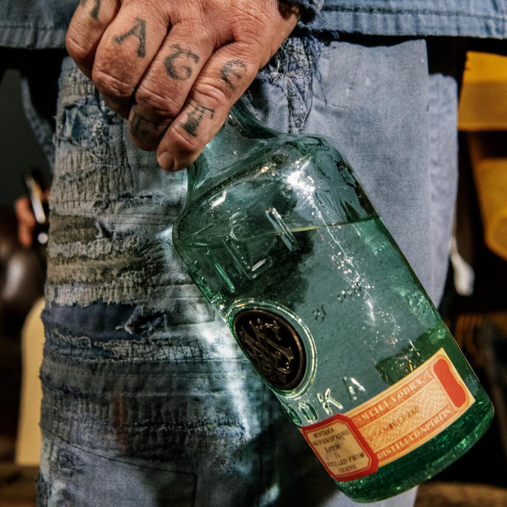 Person holding a Meilli vodka bottle with a label, wearing a denim jacket and pants.