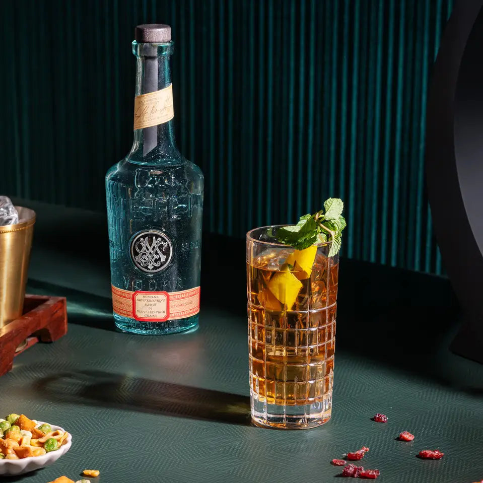 Load image into Gallery viewer, A bottle of Meili Vodka with a dark cap and orange label stands beside a tall textured tumbler filled with an amber drink, garnished with mint and a yellow citrus slice, on a dark green tabletop with scattered snacks and a dark teal background.