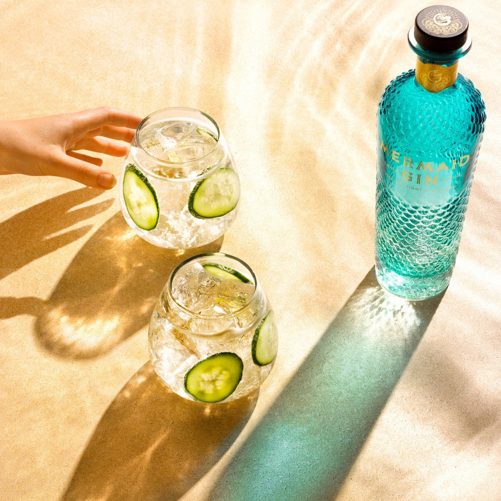 Bottle of Mermaid Gin with two glasses of gin and tonic on a sandy surface
